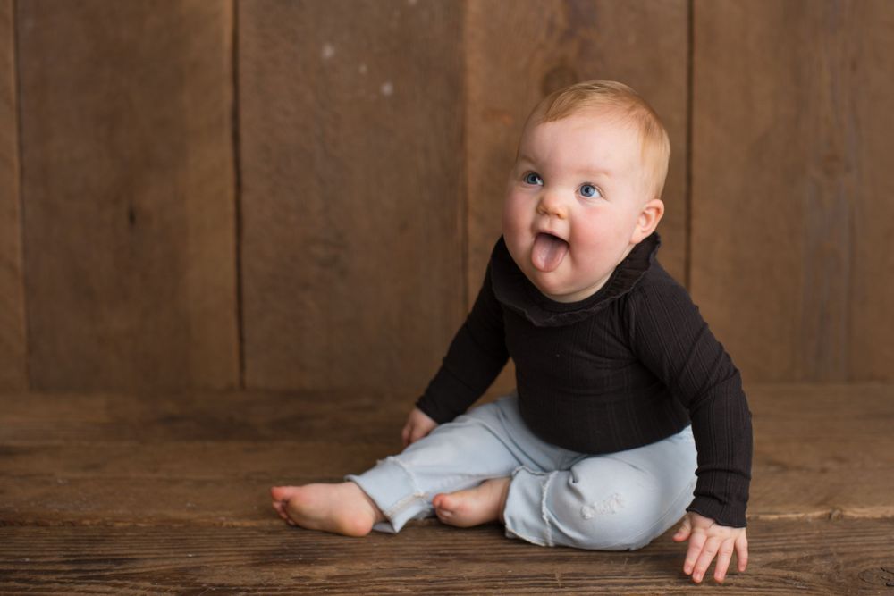 Baby in black top and gray pants sits playfully on wooden floor.