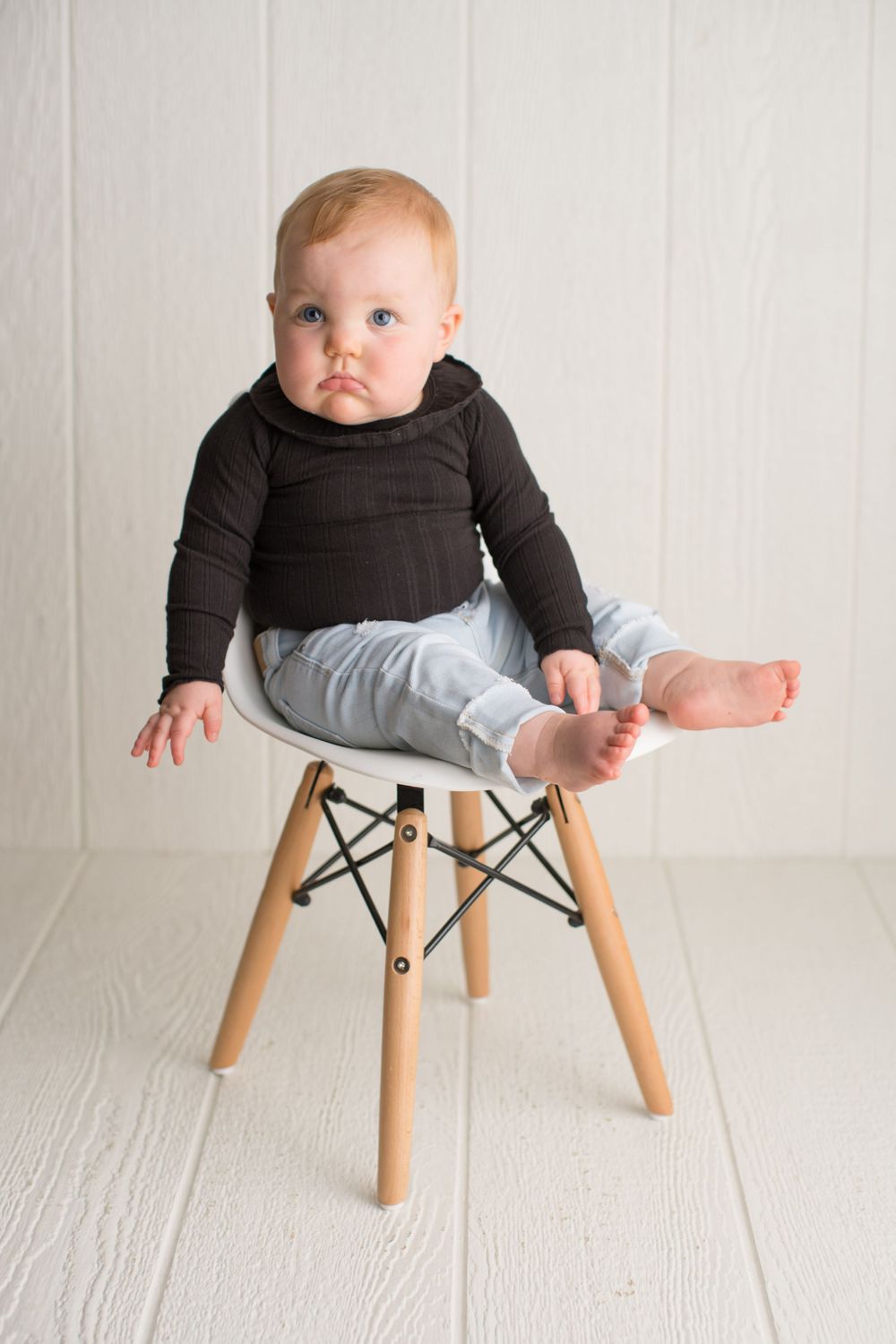 Baby in black sweater and gray pants sits on modern white chair against white background.