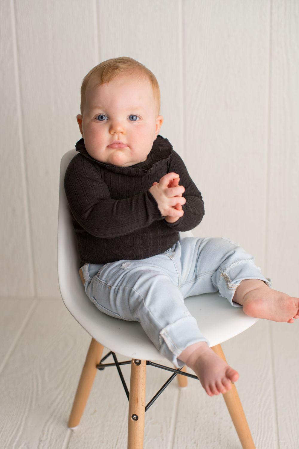Baby wearing dark hoodie and light pants sits on modern white chair for portrait.