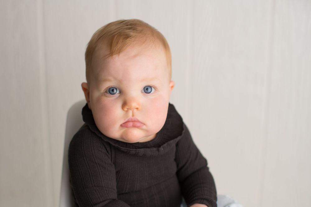 Baby in dark hoodie sits against white wall for professional portrait.
