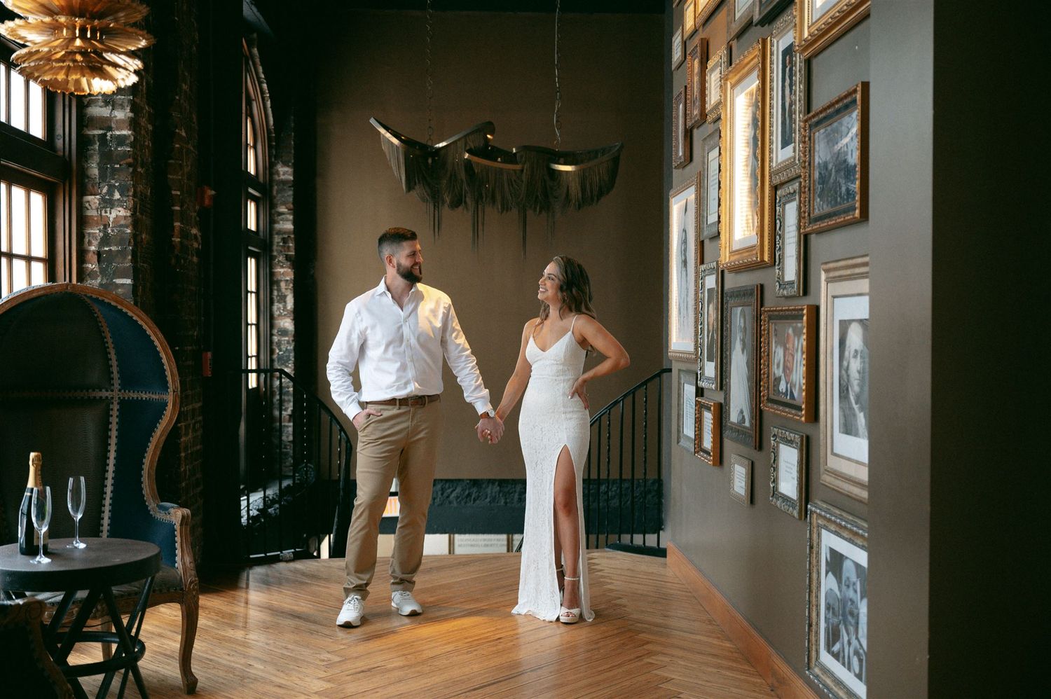 A couple holds hands in an elegantly decorated room with vintage decor, wooden floors and framed photos on the wall.