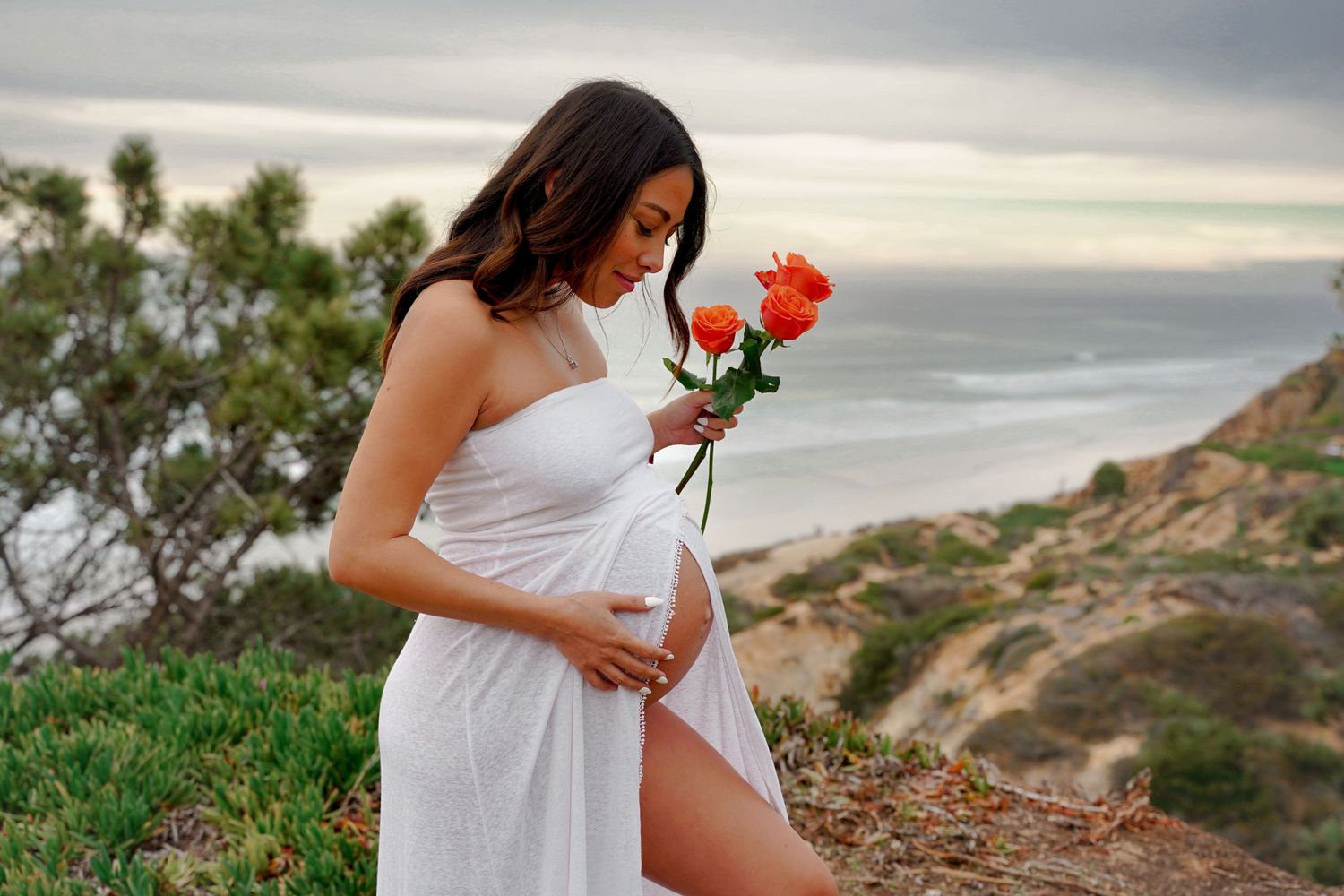 Figure in flowing white dress holds red roses against coastal cliff backdrop.