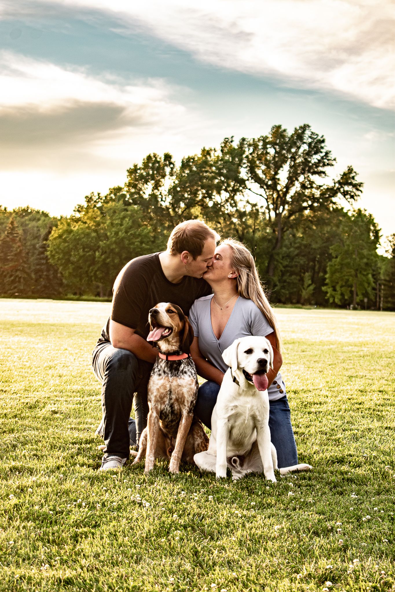 A couple and their two dogs pose together in a sunny meadow at golden hour.