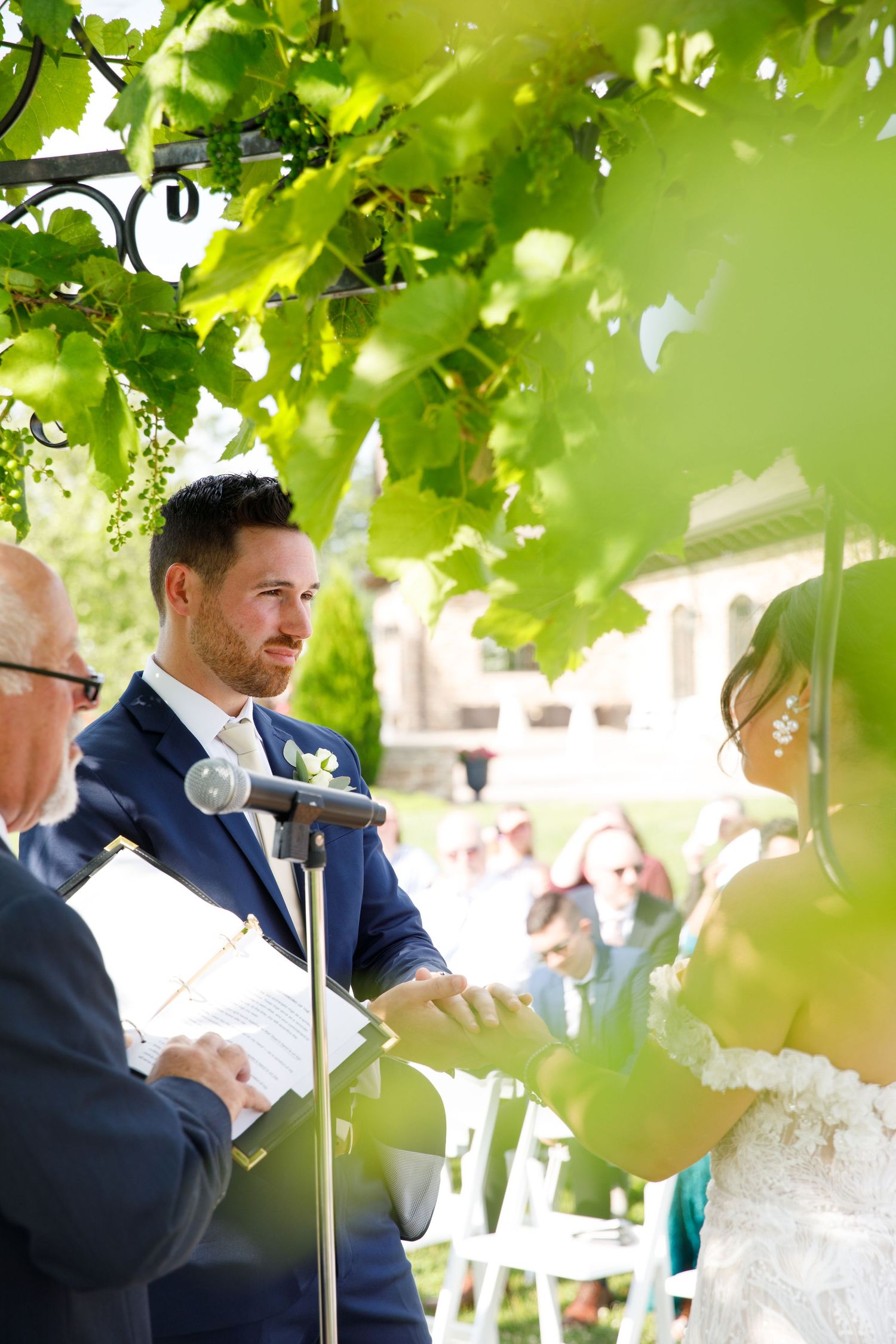 Groom in a navy suit gazing at his bride during an outdoor wedding ceremony beneath lush green vines.