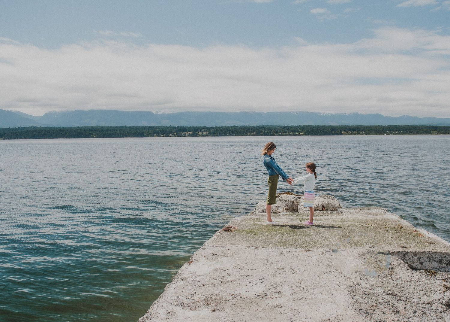 Two people stand on a concrete pier overlooking a serene lake with mountains in the distance.
