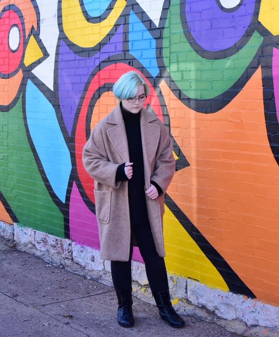 Person in beige coat against colorful street art mural featuring bold geometric shapes.