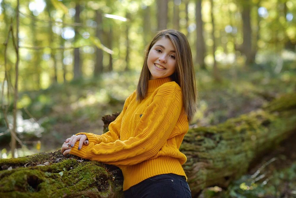 Person in bright yellow sweater sitting on moss-covered log in sunlit forest setting.