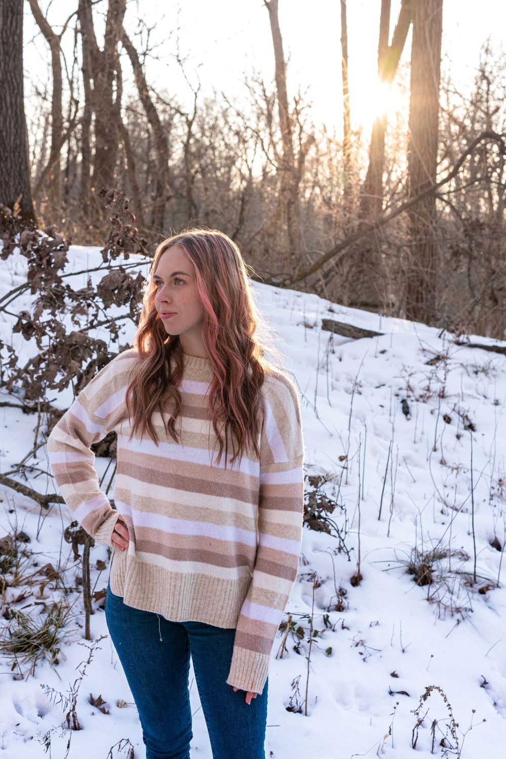 A person in a striped sweater poses in a snowy wooded area during golden hour.