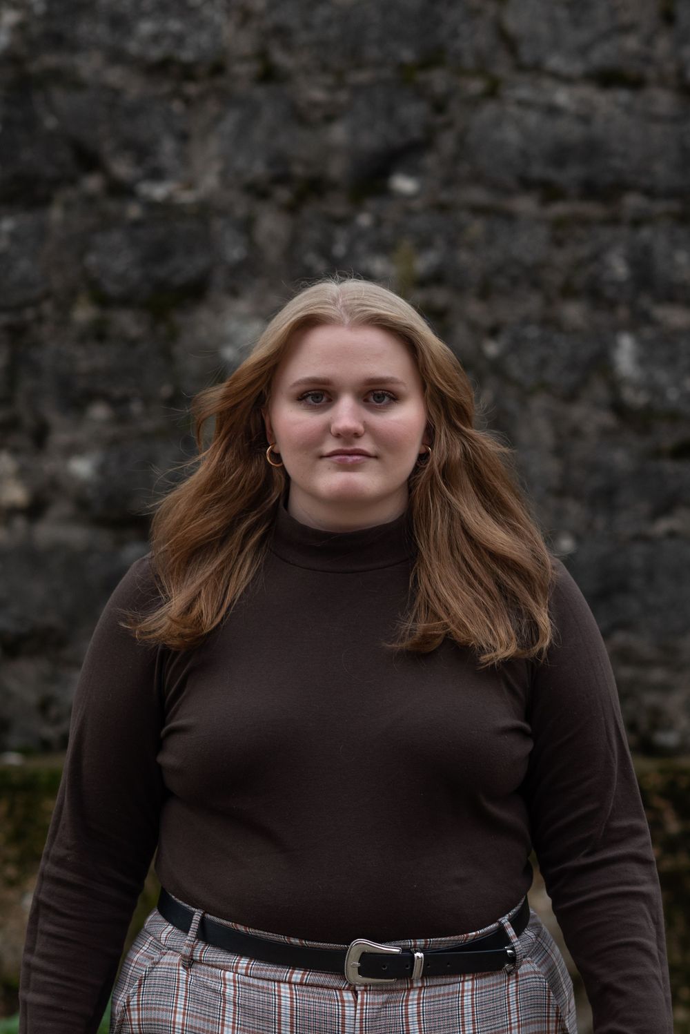 Portrait against stone wall in brown turtleneck and plaid pants.