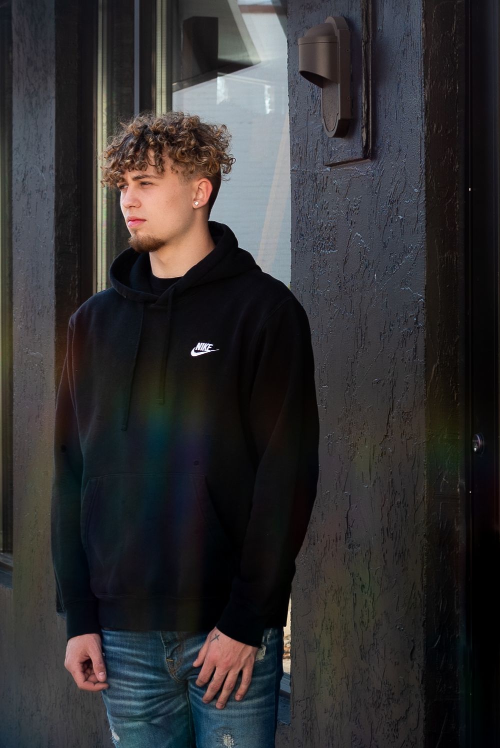 A person in a black Nike hoodie stands against a dark wall with a dramatic lighting effect.