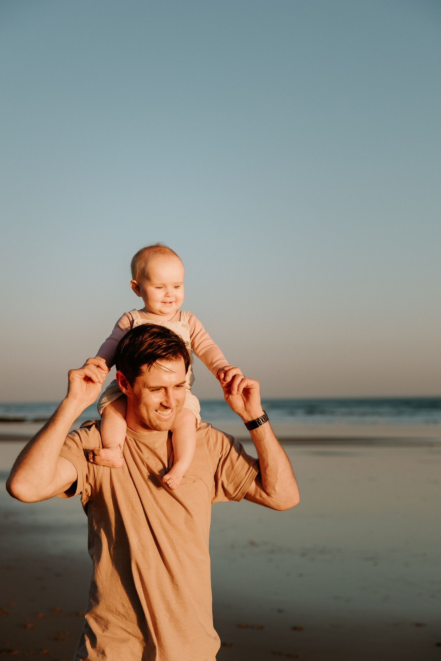 Parent carrying child on shoulders at beach during golden hour.