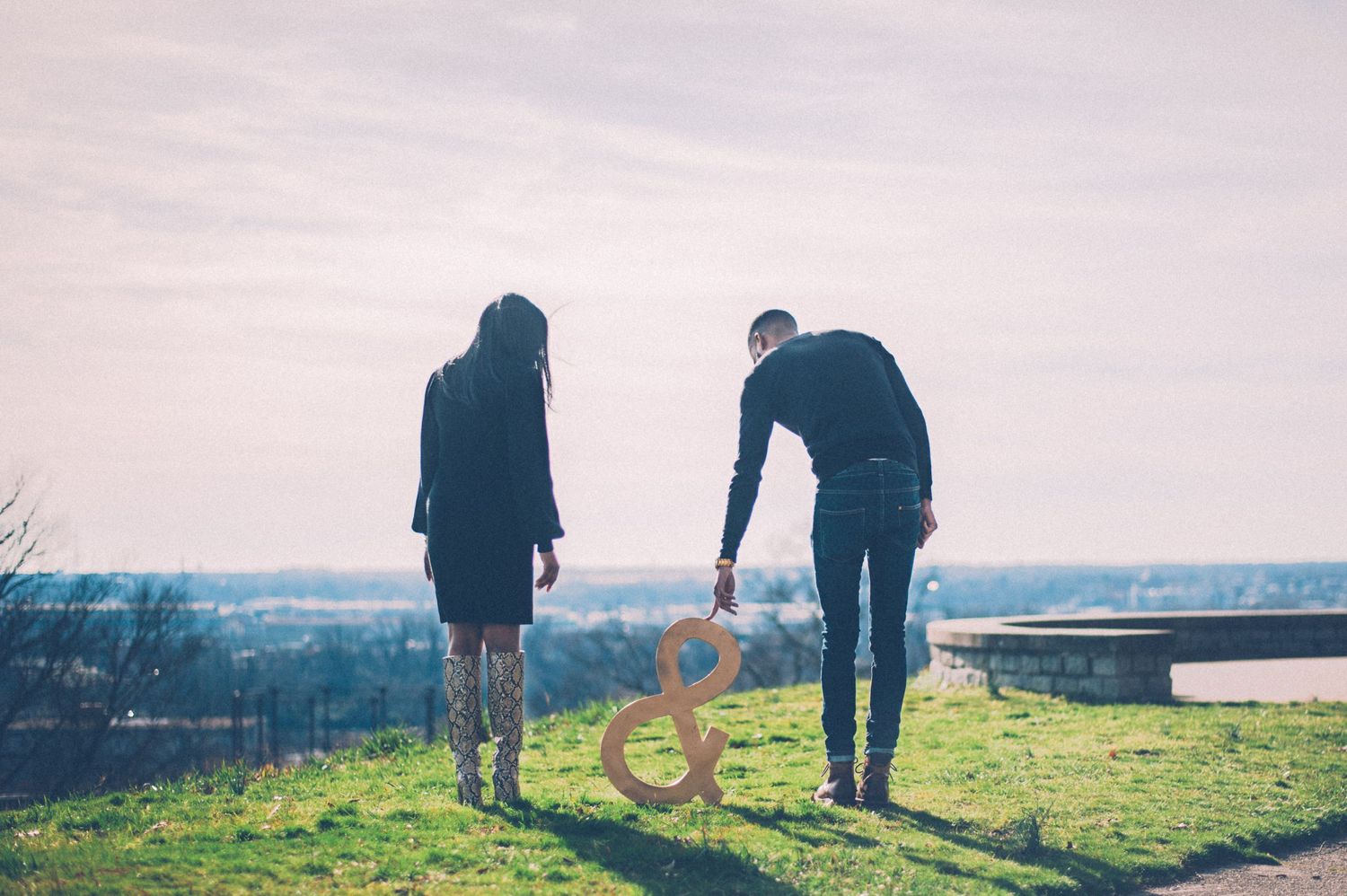 Two people standing on hilltop holding ampersand sign against scenic view.