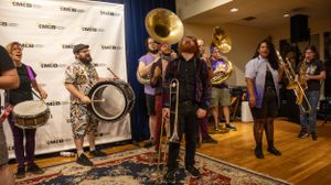 Musicians with brass instruments and drums perform at event with backdrop.