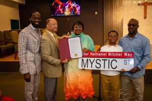 Group presents ceremonial street sign reading 'Mystic St' during indoor ceremony.