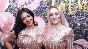 Two women in sparkly pink dresses pose at party with balloons and glitter backdrop.
