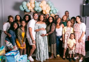A large group celebrates at a baby shower with blue and peach balloons decorating the festive indoor space.