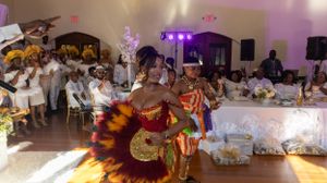 Traditional dancers perform in colorful costumes at a formal event with white-dressed attendees watching.