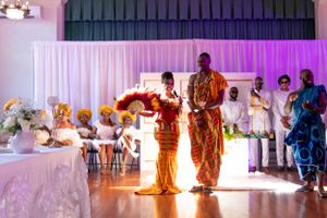 Two people in vibrant orange traditional clothing dance together at an indoor celebration.