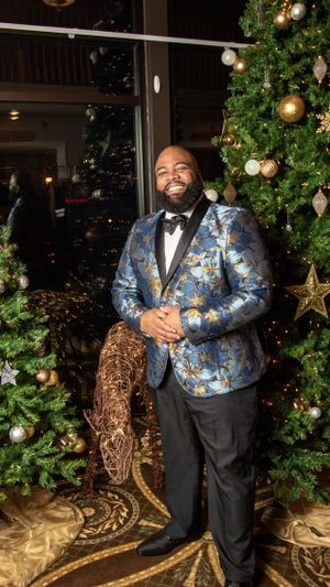 Person in blue floral tuxedo jacket posing by decorated Christmas tree.