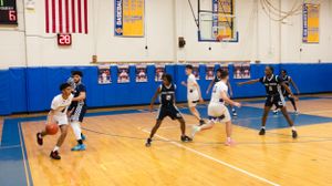 High school basketball teams compete on court with American flag visible in background.