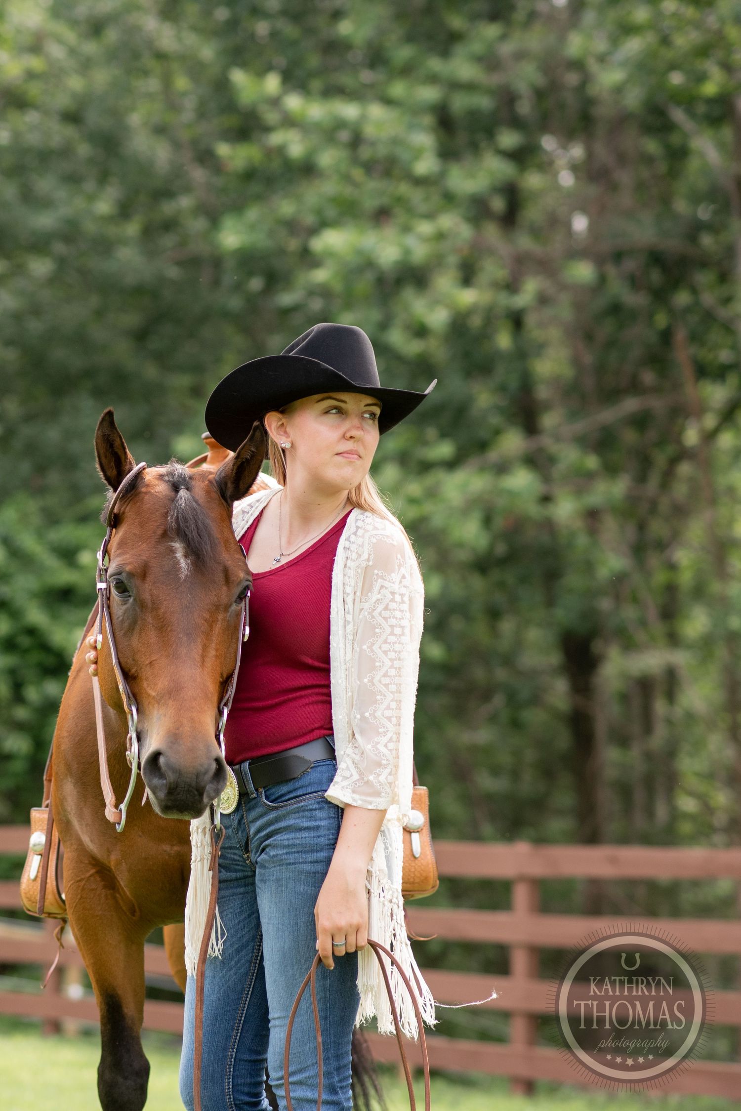 Someone in western wear and white cardigan poses with their brown horse by a fence.