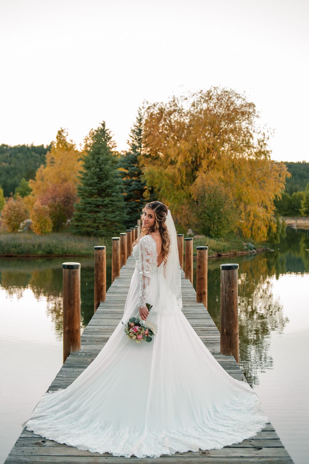 Bride in flowing white dress posing on wooden dock with autumn trees in background.
