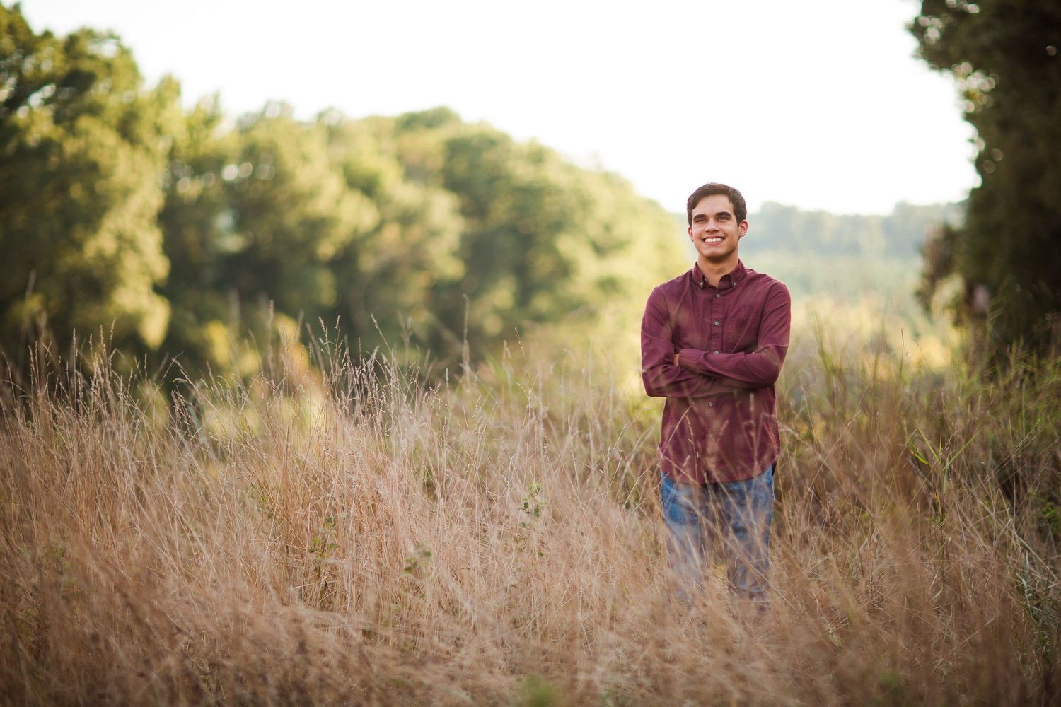 Portrait in tall grass field with mountains and trees in background during golden hour.
