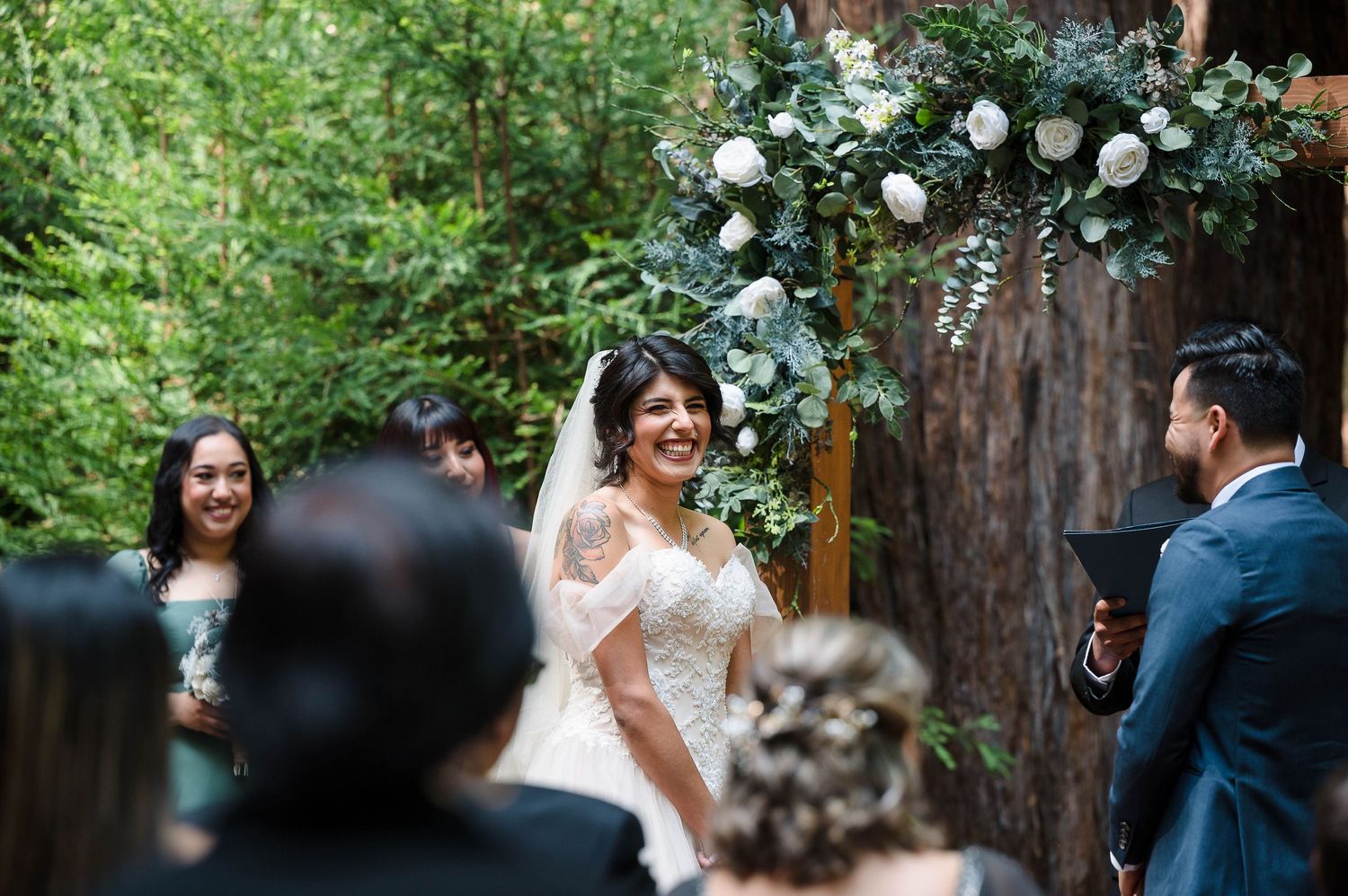 A joyful outdoor wedding ceremony takes place under towering redwood trees decorated with white floral arrangements.