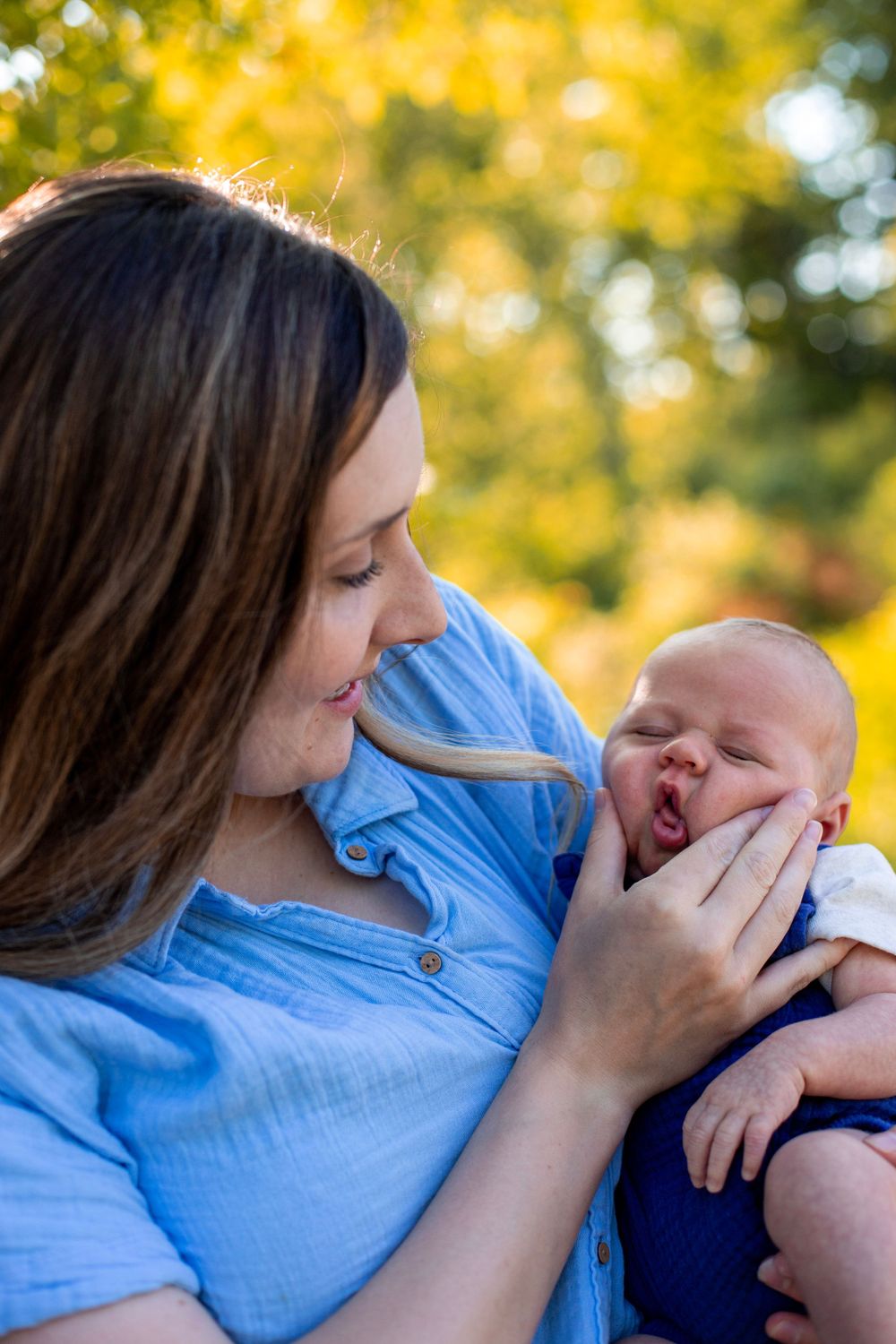 Tender moment between mother and newborn baby with golden sunlit trees in background.