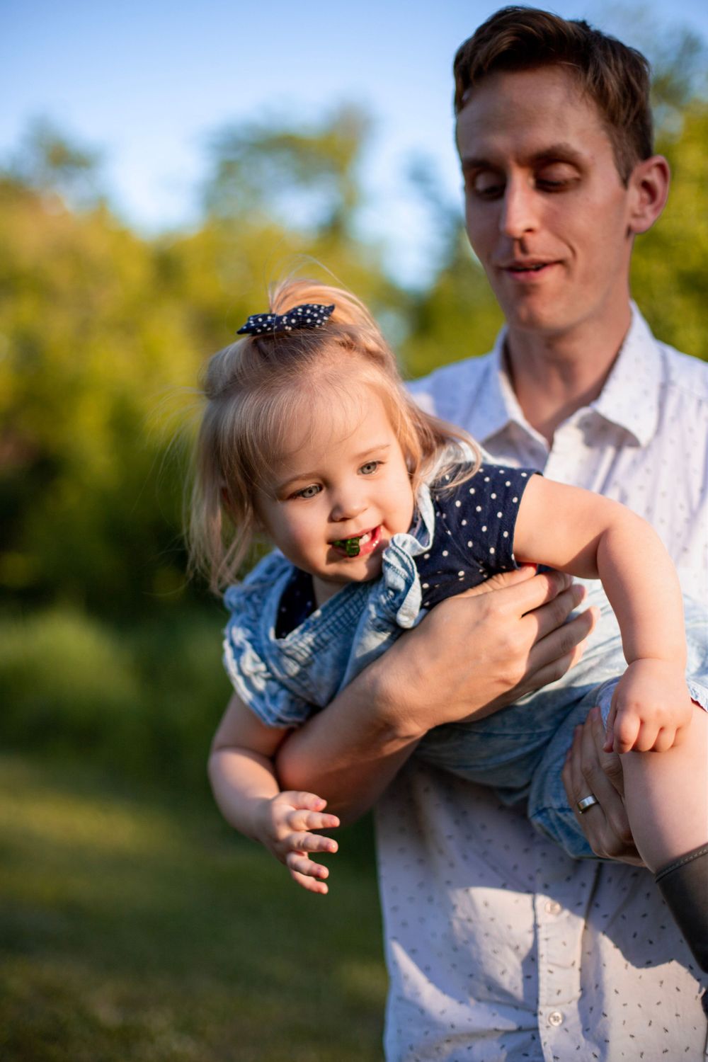 A joyful toddler in a polka dot shirt and denim outfit laughs while being held up during a sunny outdoor photoshoot.