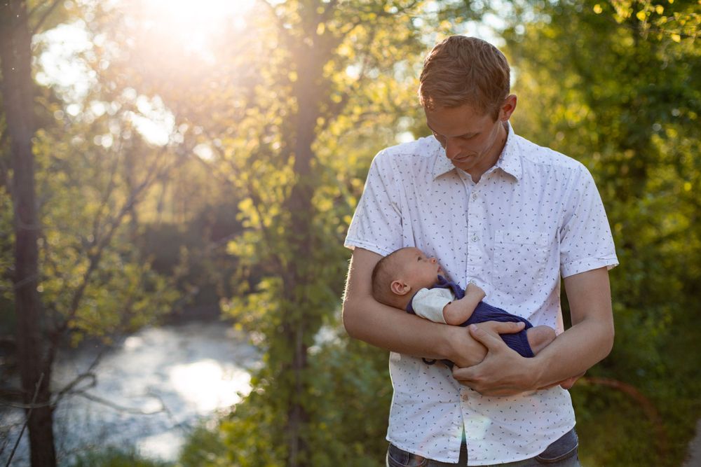 Adult holding a swaddled newborn baby outdoors at sunset with sunlit trees in the background.