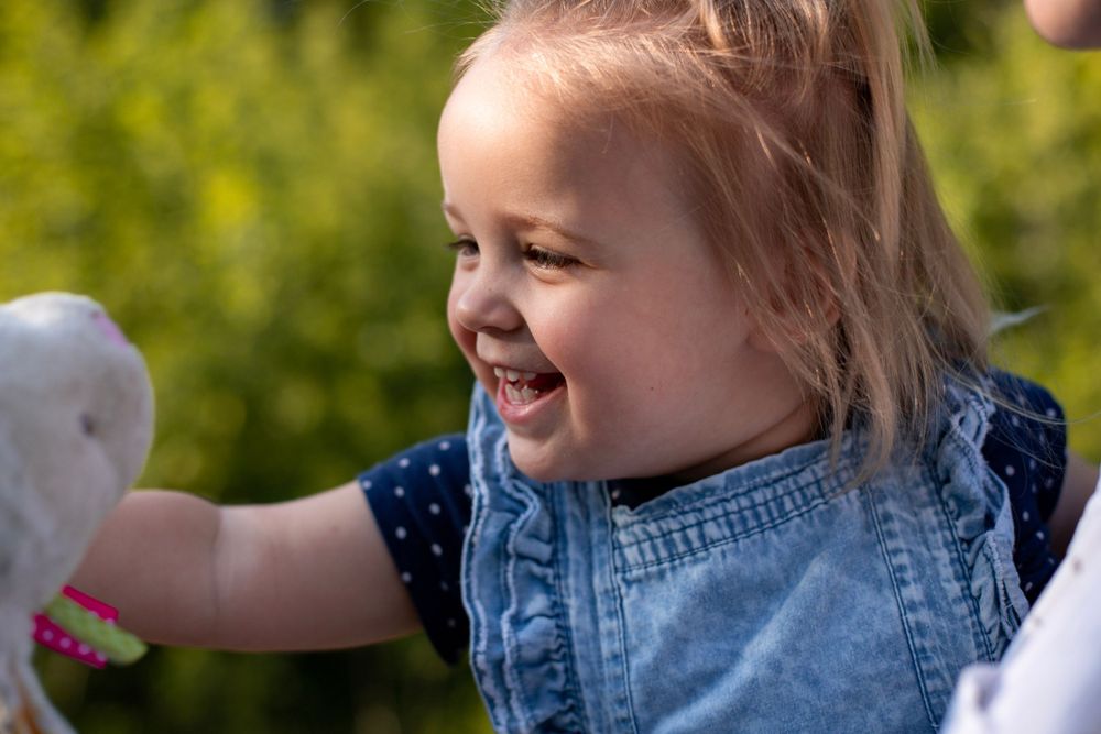 Close up of laughing toddler wearing denim and polka dots against green foliage background.