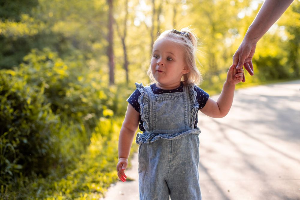 Toddler in denim overalls walking while holding hand on sunny nature path.
