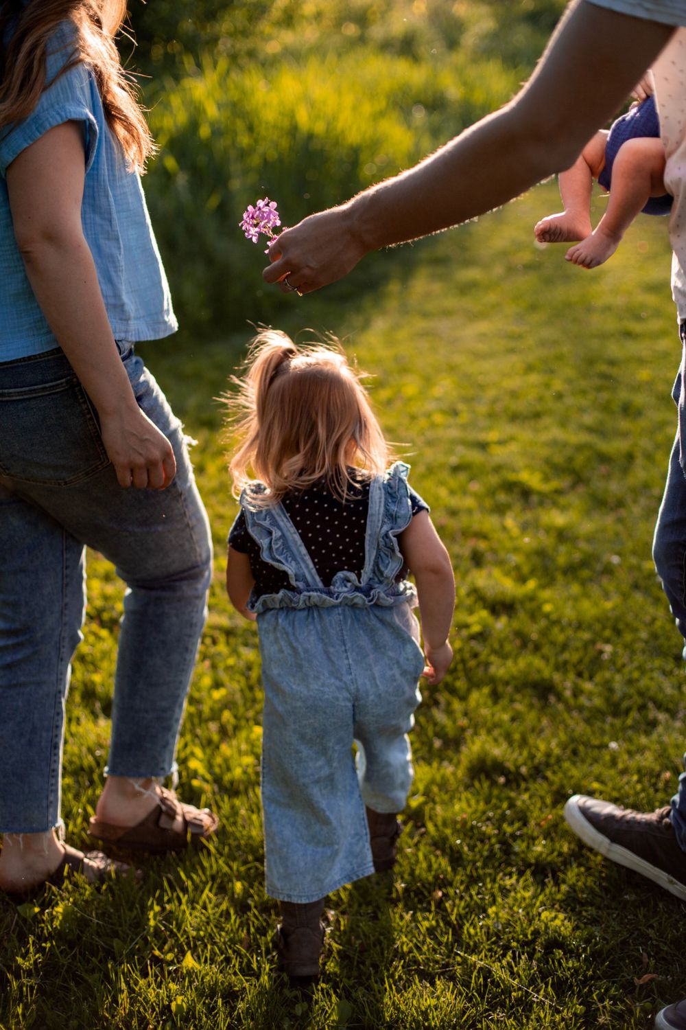 A small child in denim overalls reaches for a flower being offered in a sunny outdoor setting.