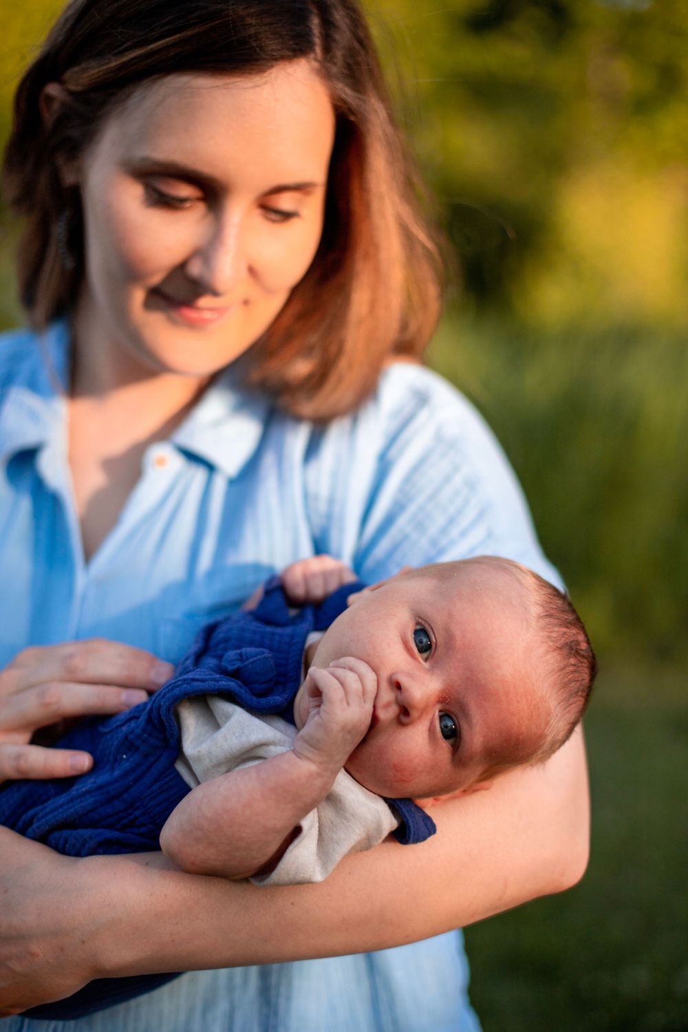 A parent tenderly holds a small baby wearing blue while standing in warm evening light outdoors.