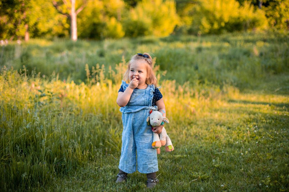 A laughing toddler in denim dress holds a stuffed toy while standing in a sunlit meadow.