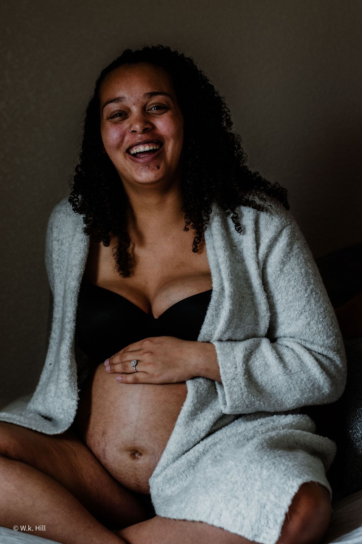 An expectant mother in a cozy gray cardigan smiles warmly during a maternity photoshoot.