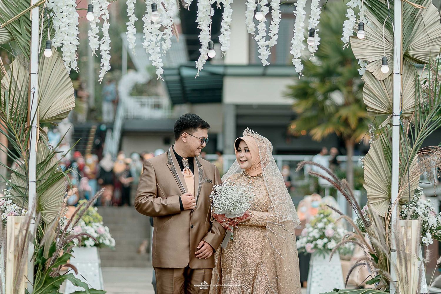 Couple shares intimate moment under floral wedding arch decorated with hanging white blooms.