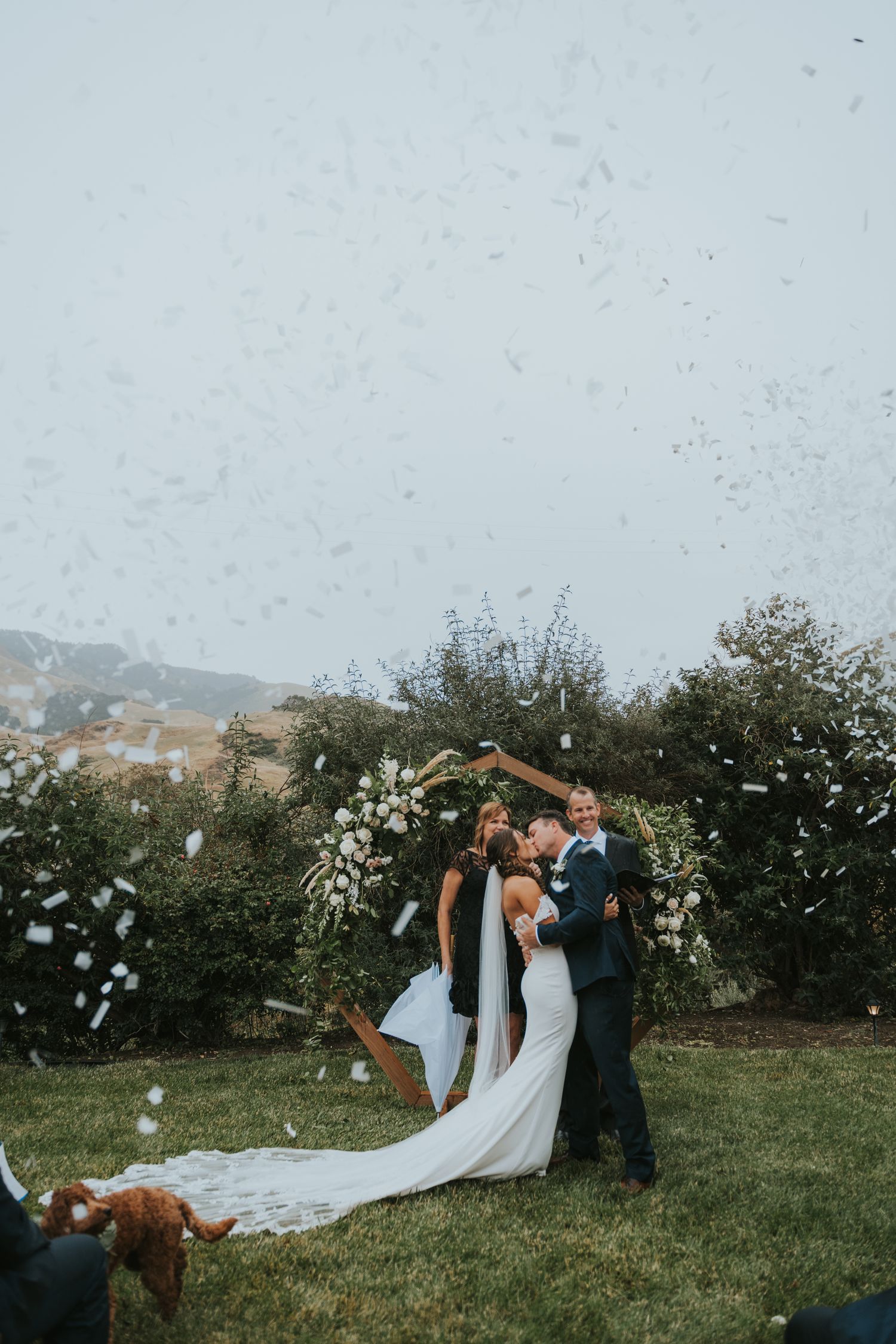 A couple kisses under a floral arch with confetti during a stunning destination wedding in San Luis Obispo, CA