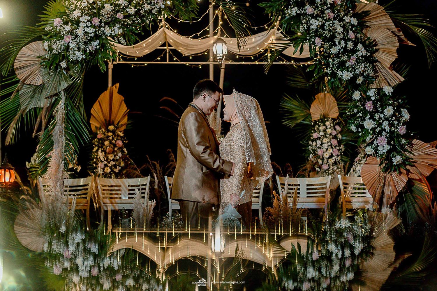 Evening wedding ceremony setup with floral arch and hanging lanterns creating reflection.
