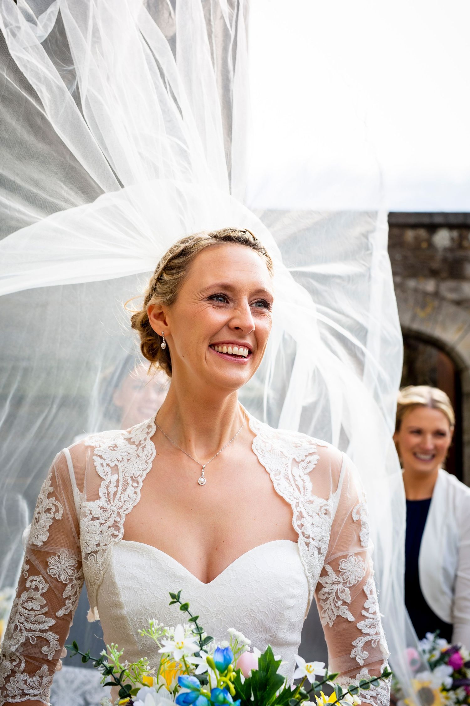 A bride wearing a lace wedding dress and flowing veil smiles while holding a colorful bouquet.