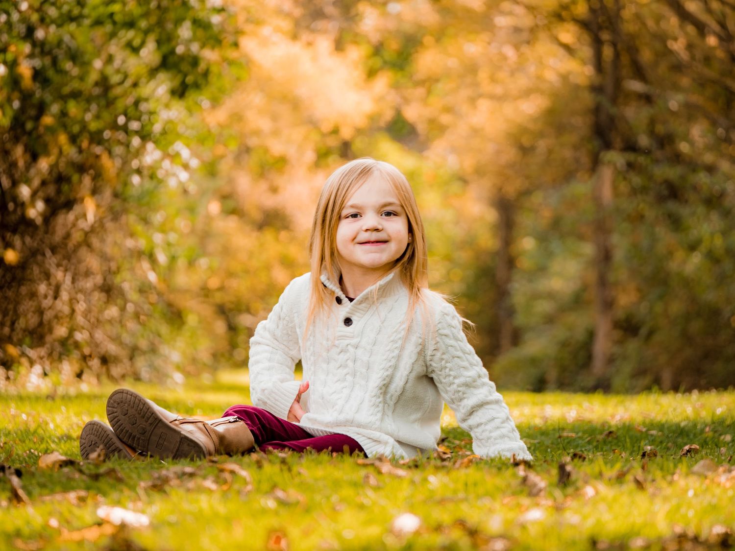 Child in white sweater sits on autumn leaves with bright fall foliage in background.