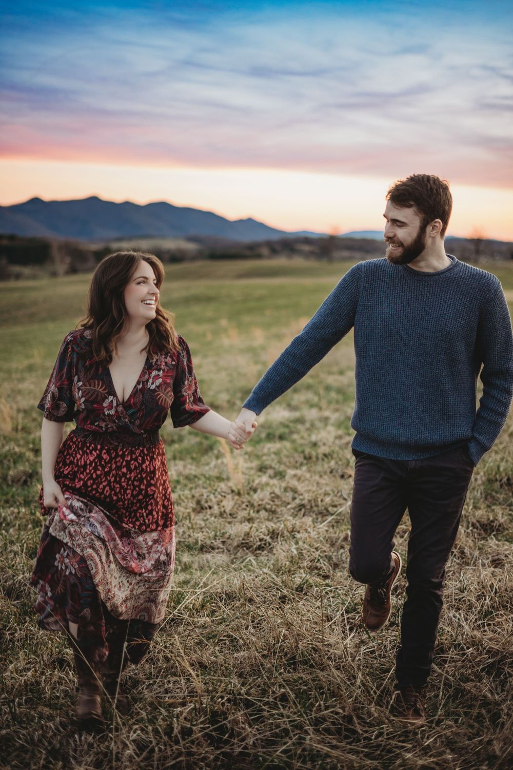 A couple walks hand in hand through a grassy meadow with mountains and sunset in the background.
