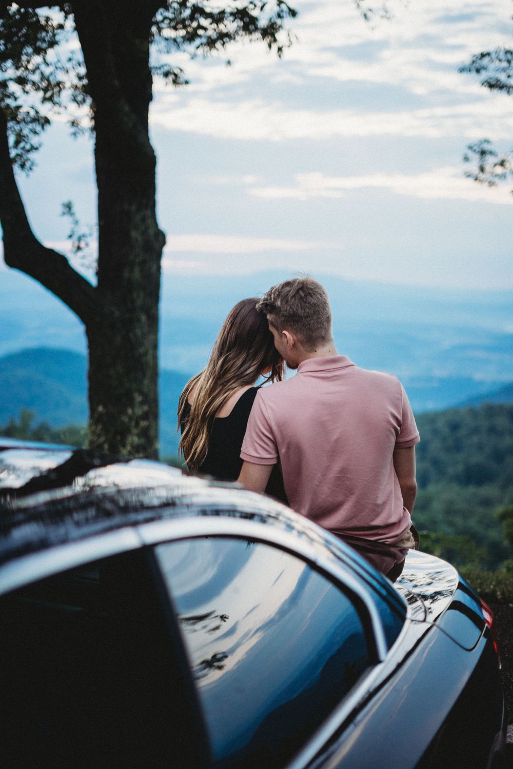 Couple sits together on their car overlooking a scenic mountain landscape.