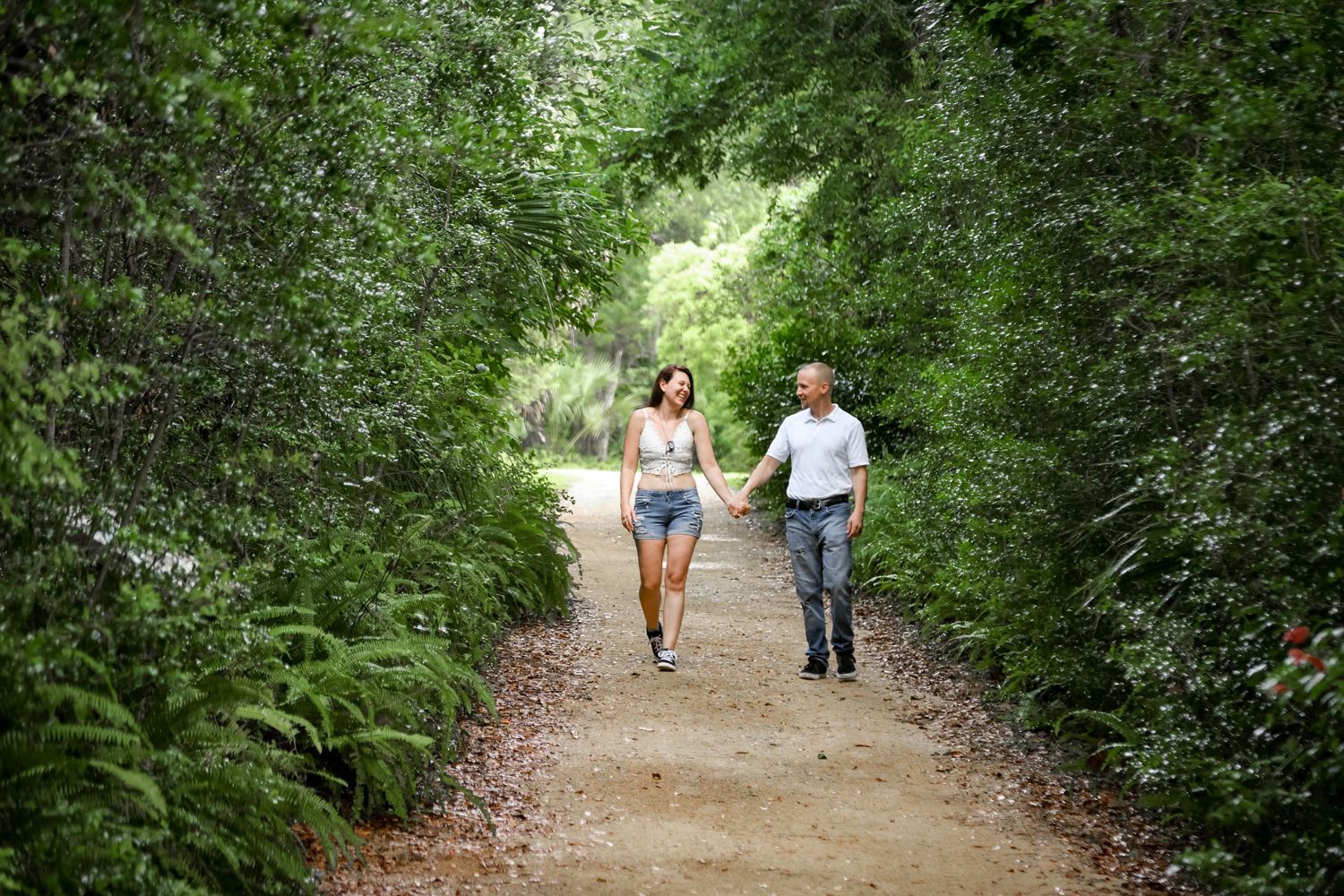 Two people jog together on a tree-lined dirt path through lush green foliage on a bright summer day.