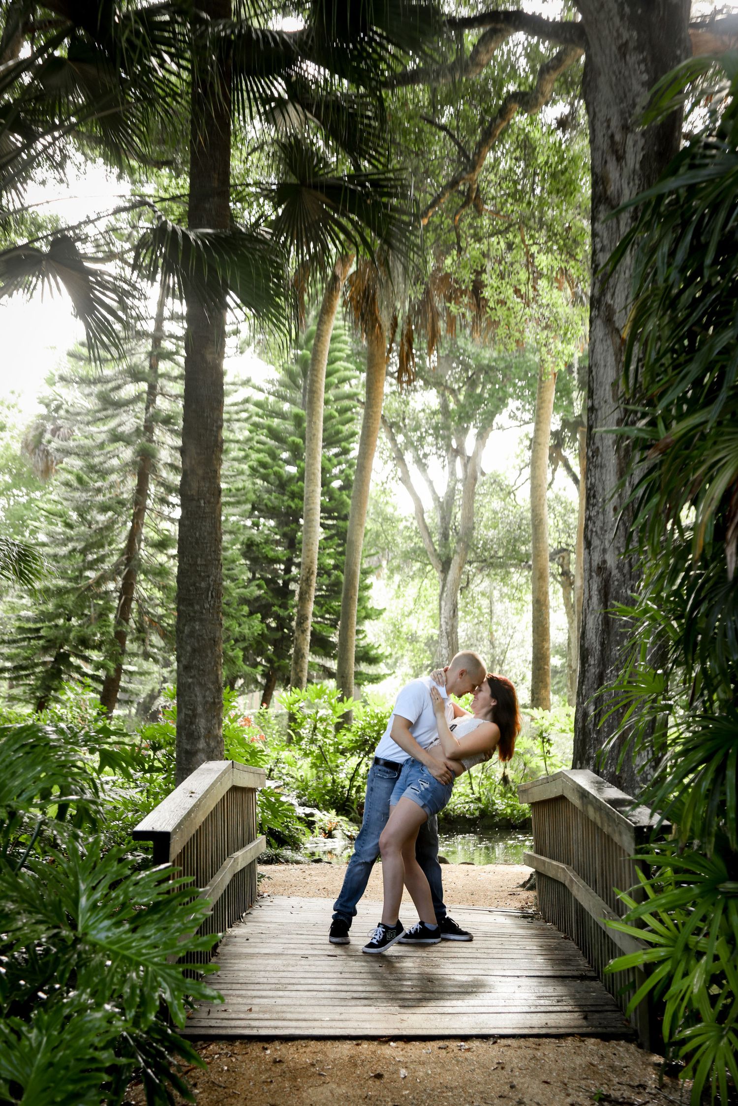 A couple shares a romantic moment on a wooden bridge surrounded by palm trees and Spanish moss.