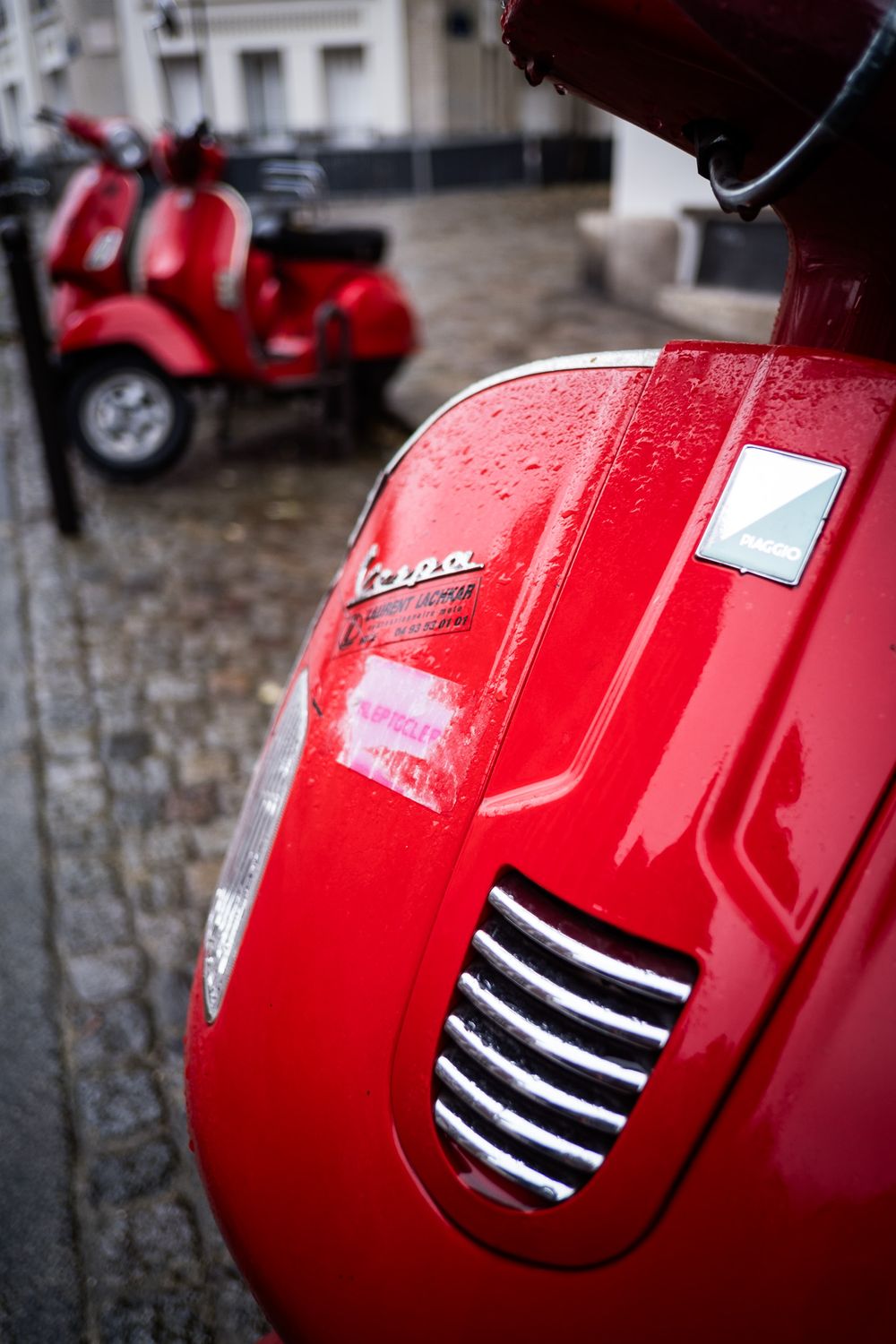 Close-up of a shiny red Vespa scooter with raindrops on cobblestone street in European city.