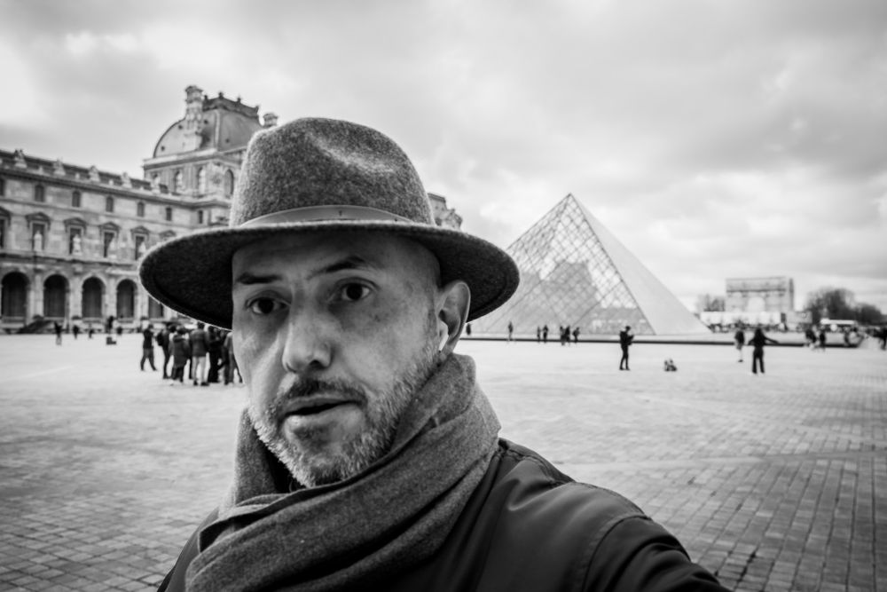 Man in fedora hat taking selfie at Louvre Museum glass pyramid Paris black and white.
