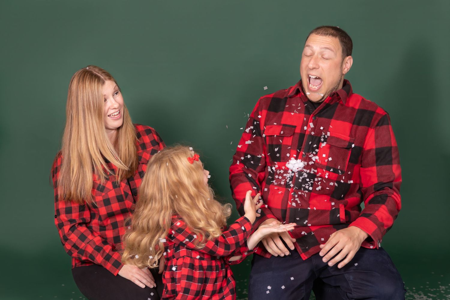 family wearing red plaid on green backdrop child is throwing fake snow at dad everyone laughing studio clayton candid