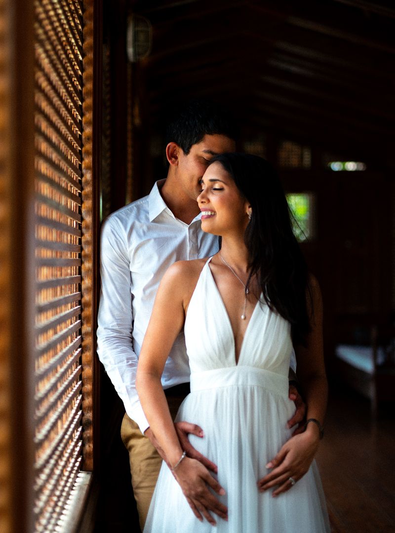 A loving couple shares an intimate moment by wooden window blinds in soft natural lighting.